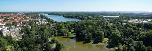 Luftaufnahme eines Parks mit einem Teich und vielen Bäumen, umgeben von einer Stadtlandschaft.