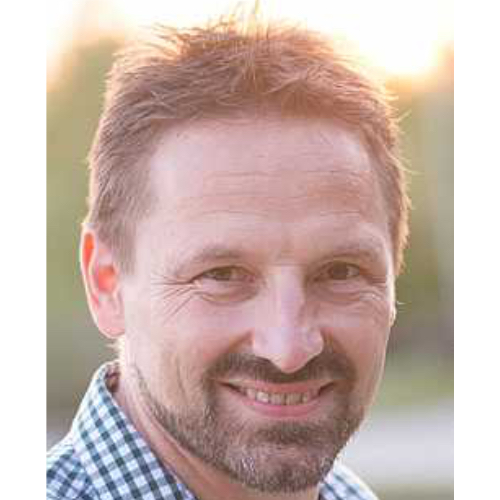 Mann mit Bart und kurzem Haar, der in die Kamera lächelt, trägt ein kariertes Hemd.