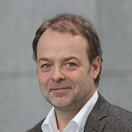 Porträt eines Mannes mit kurzen, braunen Haaren und blauen Augen in einem grauen Anzug.