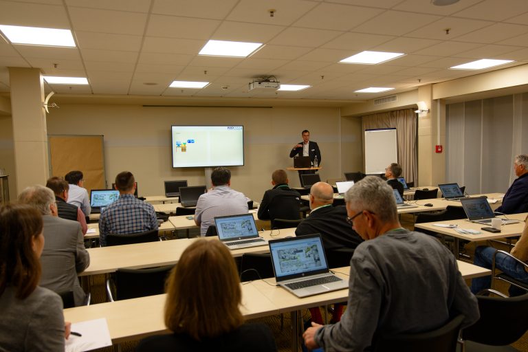 Ein Referent steht vor einem Bildschirm und präsentiert vor einem Publikum in einem Konferenzraum.