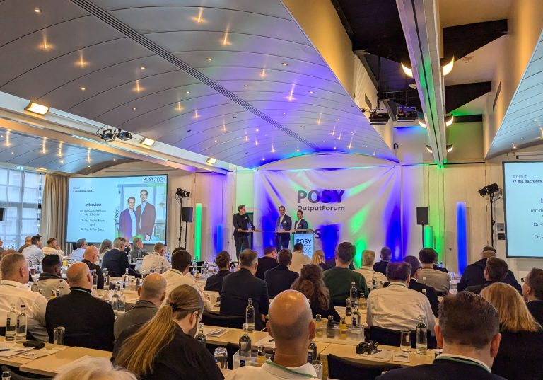 Zwei Redner auf einer Bühne während des POSY OutputForum 2024 mit Publikum im Vordergrund.